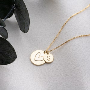 May include: Gold necklace with two charms. One charm is a circle with a heart cutout and the other charm is a circle with the letter "S" engraved on it.