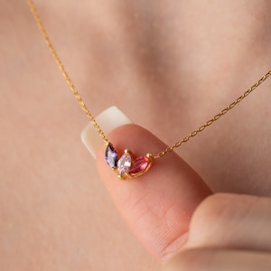May include: A gold chain necklace with a delicate pendant featuring three small, colourful gemstones. The pendant is shaped like a cluster of leaves, with one purple, one pink, and one clear gemstone.