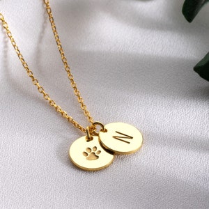 May include: Gold necklace with two charms. One charm is a paw print and the other charm is a circle with the letter N.