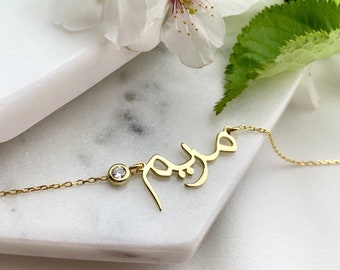 Puede incluir: Un collar de oro con una cadena delicada y un colgante con un nombre escrito en escritura árabe con un pequeño strass transparente.