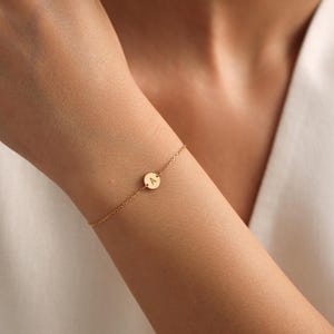 Pode incluir: Um bracelete de corrente dourada com um pequeno charme redondo com a letra 'A'.