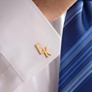May include: A gold-toned metal cufflink featuring the letters E and K joined together, attached to the white fabric cuff of a long-sleeved shirt. The cufflink has a polished finish and is positioned next to a blue and light blue striped necktie.