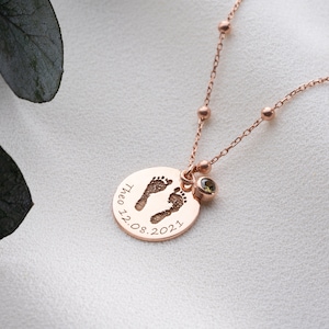 Op de afbeelding: Een roze gouden ketting met een ronde hanger met een babyvoetje en de datum "12.08.2021". De hanger is bevestigd aan een delicate ketting met kleine kralen. Een kleine groene edelsteen is aan de hanger bevestigd.