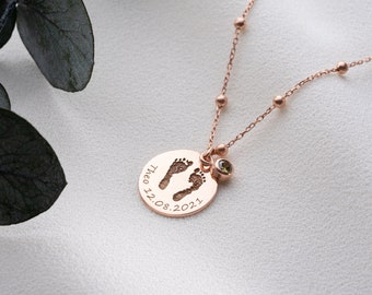 May include: A rose gold necklace with a round pendant featuring a baby's footprint and the date "12.08.2021". The pendant is attached to a delicate chain with small beads. A small green gemstone is attached to the pendant.