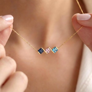 May include: A gold necklace with three square gemstones in navy, pink, and light blue. The gemstones are set in gold bezels and are connected by a delicate gold chain. The necklace is being held up by a person.