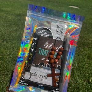 May include: A clear plastic bag filled with religious themed items, including a wooden cross, a brown rosary, a black keychain with the text "John 3:16", a black keychain with the text "Psalm 37:4", a black and white sticker with the text "Walk by faith", and a black and white sticker with the text "Let that be a motivation will you and will".