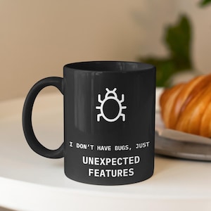 May include: A black coffee mug with a white bug icon and the text "I don't have bugs, just unexpected features".