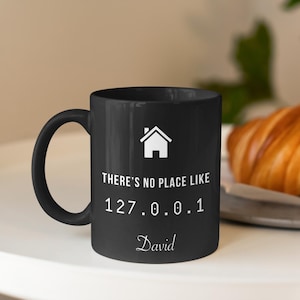 May include: Black coffee mug with white text that reads "There's no place like 127.0.0.1 David". The mug has a white house icon and a black handle.