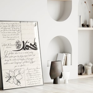 May include: A black and white Islamic calligraphy print with a floral design. The print features Arabic text and a quote from the Prophet Muhammad. The print is framed in a black frame and is hanging on a white wall.