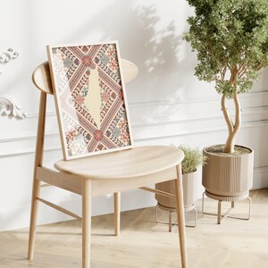 May include: A framed print of a stylized map of Palestine with a traditional Palestinian embroidery pattern in red, blue, and white on a beige background. The print is hanging on a light wood chair in a room with white walls and a potted plant.