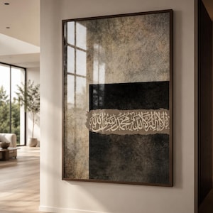 May include: A framed abstract artwork with a textured background in shades of grey, brown, and black. The artwork features Arabic calligraphy in white and a dark brown wooden frame. The piece is displayed in a well-lit room.