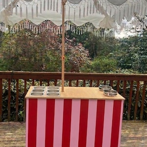 May include: A striped red and pink ice cream cart with a wooden countertop and stainless steel containers. The cart has a white canopy with fringe and is set on a wooden deck. The cart is designed for outdoor use.