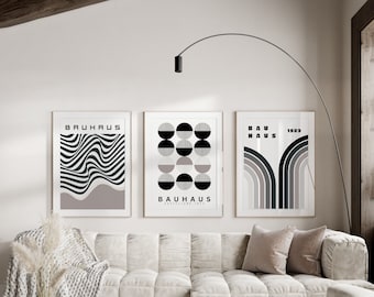 May include: Three minimalist black and white geometric art prints with the text "Bauhaus" and "1923" on a white wall above a white couch with beige pillows.