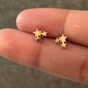 Puede incluir: Un par de pendientes de botón dorados con forma de estrella. Cada pendiente presenta un grupo de tres estrellas de diferentes tamaños. Los pendientes son pequeños y delicados, adecuados para el uso diario. Los pendientes se muestran en un dedo.