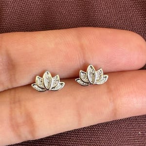 Puede incluir: Un par de pendientes de botón plateados con forma de flor de loto. Cada pendiente presenta un diseño texturizado de múltiples pétalos con un acabado metálico pulido, capturando la forma simétrica de una flor en un estilo compacto.