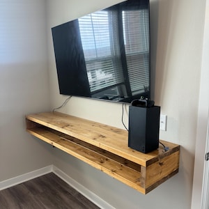 May include: A wooden floating shelf with a lower shelf, mounted on a wall, with a black TV and a black gaming console on top.