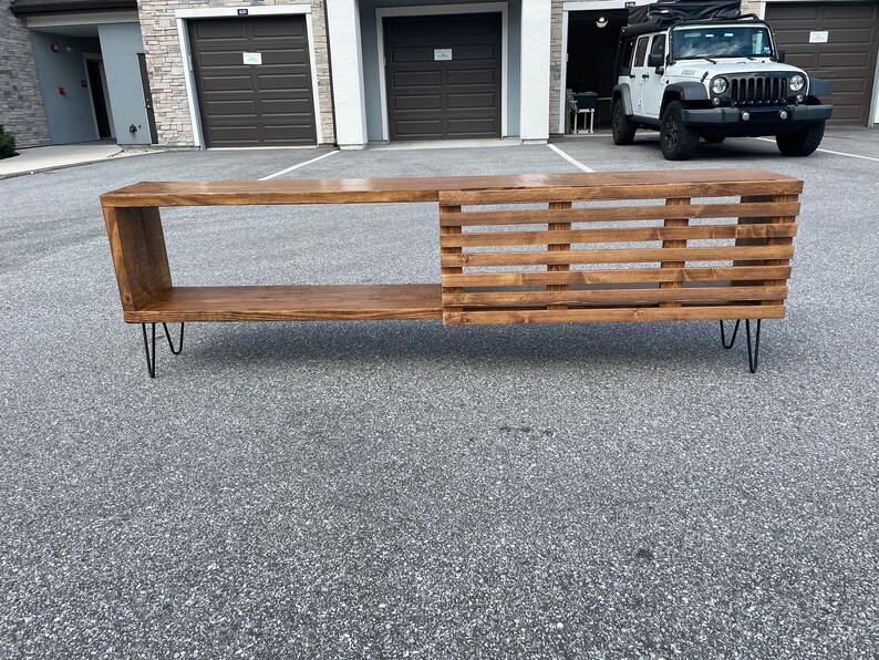 May include: A wooden media console with a slatted design on one side and a shelf on the other. The console is supported by hairpin legs and has a dark brown stain.