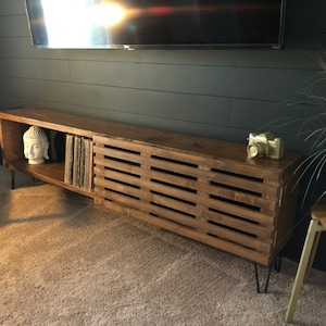 May include: A long, dark brown wooden entertainment center with a slatted door on the right side and open shelves on the left. The shelves hold a white Buddha statue and vinyl records. The entertainment center has black hairpin legs.