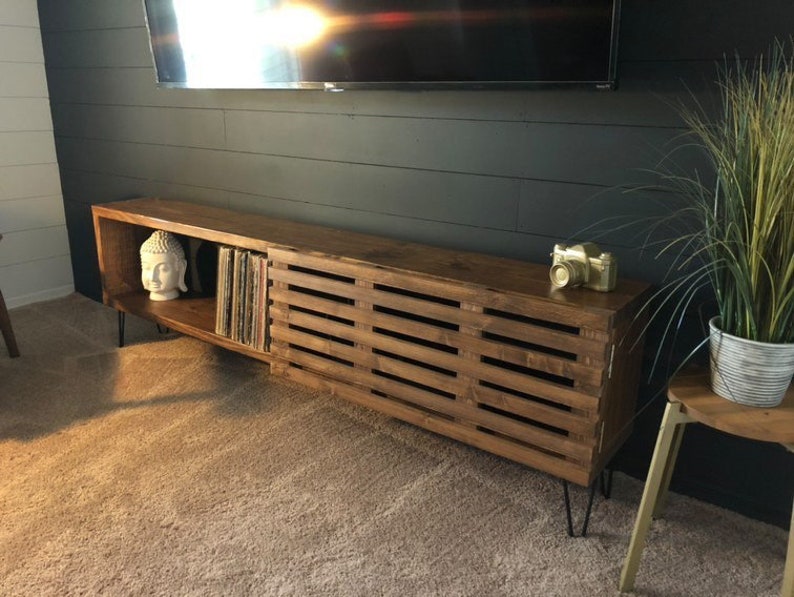 May include: A long, dark brown wooden entertainment center with a slatted door on the right side and open shelves on the left. The shelves hold a white Buddha statue and vinyl records. The entertainment center has black hairpin legs.