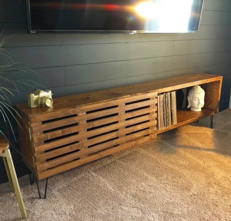 May include: A long, wooden entertainment center with a slatted door on the left and open shelves on the right. The shelves are filled with vinyl records and a white Buddha statue. The entertainment center is on a carpeted floor and has black hairpin legs.