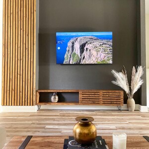 May include: A dark brown wooden media console with a slatted design and black hairpin legs. The console is against a dark gray wall with a flat screen TV mounted above it. There is a vase with dried pampas grass on the right side of the console and a gold vase on the coffee table in front of the console.
