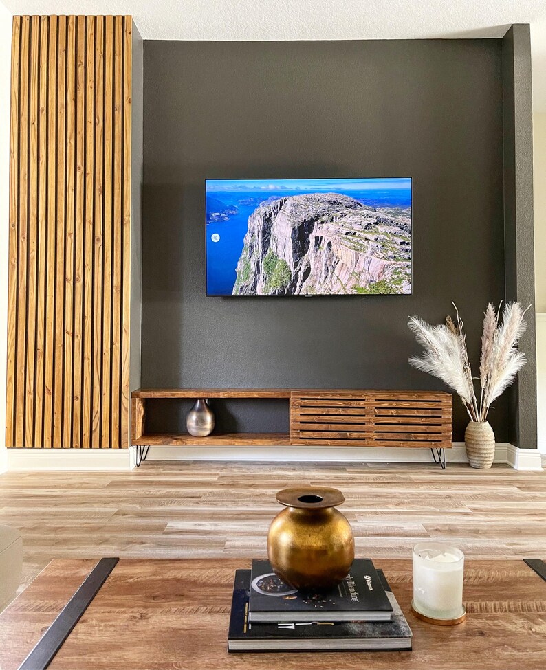 May include: A dark brown wooden media console with a slatted design and black hairpin legs. The console is against a dark gray wall with a flat screen TV mounted above it. There is a vase with dried pampas grass on the right side of the console and a gold vase on the coffee table in front of the console.