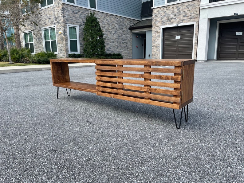 May include: A wooden media console with a slatted design and a shelf. The console is supported by black hairpin legs.