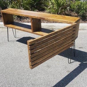 May include: A wooden entertainment center with a unique angled design. The unit features a shelf, two compartments, and a slatted back panel. It is supported by hairpin legs.