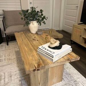 May include: A rustic wooden coffee table with a natural wood finish. The table has a wide, rectangular top and a lower shelf. There are two gold candle holders and a stack of books on the table, including a book with the word "CHANEL" on the cover.