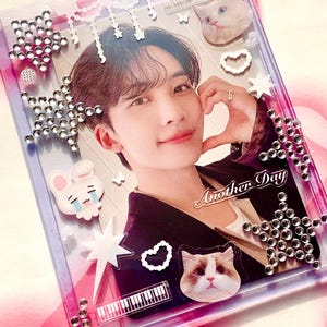 May include: A clear, rectangular photo card holder featuring a person's photo. The holder is adorned with star-shaped and round rhinestones, cat and bunny stickers, and the words "Another Day".