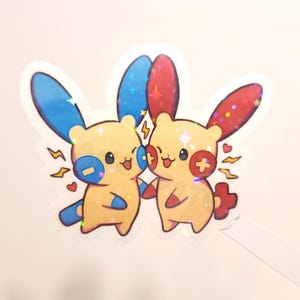May include: A holographic sticker featuring two cartoon characters with yellow bodies, blue and red ears, and smiling faces. One character has a blue plus sign, the other a red minus sign. Sparkle and lightning bolt details.