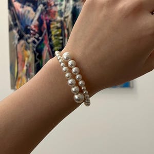 May include: Two pearl bracelets, one with large pearls and the other with smaller pearls, are worn on a wrist. The pearls are white and have a glossy finish. The bracelets are stacked together.