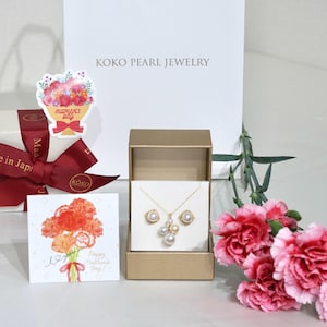 May include: A gold necklace and earrings set with pearl accents displayed in a gold box. A white gift bag with "KOKO PEARL JEWELRY" is in the background. A card with a floral bouquet and the text "Happy Mother's Day!" is also present.