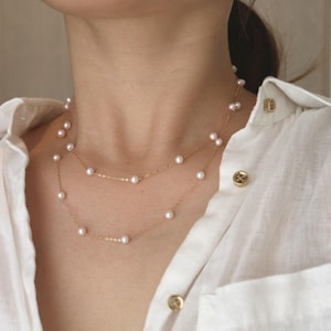 Puede incluir: Un collar de dos capas con finas cadenas de oro y pequeñas perlas redondas. El collar se lleva sobre una camisa blanca, creando un look clásico y elegante. Las perlas están espaciadas uniformemente a lo largo de las cadenas.