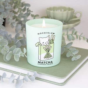 May include: A lit matcha-scented candle in a light green glass. The candle has a cartoon cat illustration and the text "MOCHIGLOW" and "MATCHA scented candle". The candle sits on a green book with eucalyptus leaves in the background.
