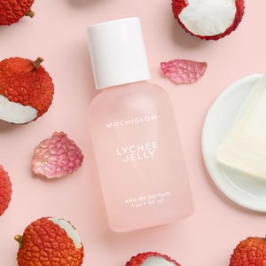 May include: A pink-tinted perfume bottle with a white cap, labeled "MOCHIGLOW LYCHEE JELLY eau de parfum 1 oz • 30 ml," is surrounded by fresh lychees and petals on a pink background. The bottle is the focal point, suggesting a fragrance product.