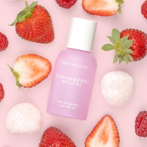May include: A light pink perfume bottle with the text "MOCHIGLOW STRAWBERRY MOCHI eau de parfum 1 oz - 30 ml" is surrounded by fresh strawberries, raspberries, and mochi on a pink background.