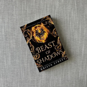 May include: A book cover with a black background and gold accents. The title is "Beast of Shadows" and the author is "USA Today Bestselling Author Krista Street". A wolf with glowing yellow eyes is featured on the cover.