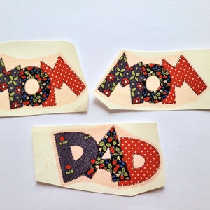 May include: Three fabric patches with the words "MOM" and "DAD" spelled out in colourful patchwork letters. The letters are made of red, blue and white fabric with floral and polka dot patterns.