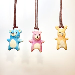 Op de afbeelding: Drie keramische hangerkettingen met dierenfiguren: een blauwe beer, een roze beer en een gele konijn. Elke hanger is geregen aan een bruin koord. De hangers hebben een glanzende afwerking en zijn met de hand beschilderd.