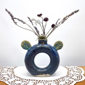 May include: A handmade, blue ceramic vase with a circular body and two leaf-shaped handles. The vase holds dried lavender and other small, dried flowers. The vase is on a white doily.