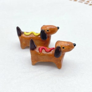 May include: Two ceramic dachshund figurines, each resembling a hot dog. The dogs are brown with black ears and noses. One has yellow mustard, the other red ketchup. The figurines are on a white surface.