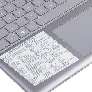 May include: A silver laptop keyboard with a white sticker displaying Visual Studio Code keyboard shortcuts. The sticker is positioned near the touchpad and includes commands for editing, navigation, and file management. The keys are gray.