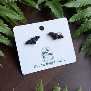 May include: Black bat-shaped stud earrings on a white card with the text "The Midnight Glen". The earrings are displayed on a dark brown surface with green foliage in the background. The earrings are a gothic or Halloween-themed accessory.