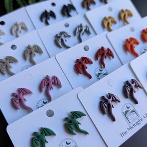 May include: A collection of dragon-shaped stud earrings in various colors, including red, purple, green, and gray, displayed on white earring cards. The earrings have detailed wing and body designs. The Midnight Glade logo is visible on some cards.