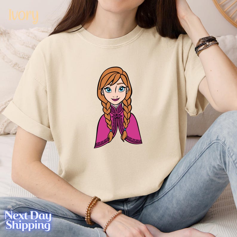 A woman wearing a white t-shirt with a cartoon illustration of a young girl with braided hair and a pink cape.