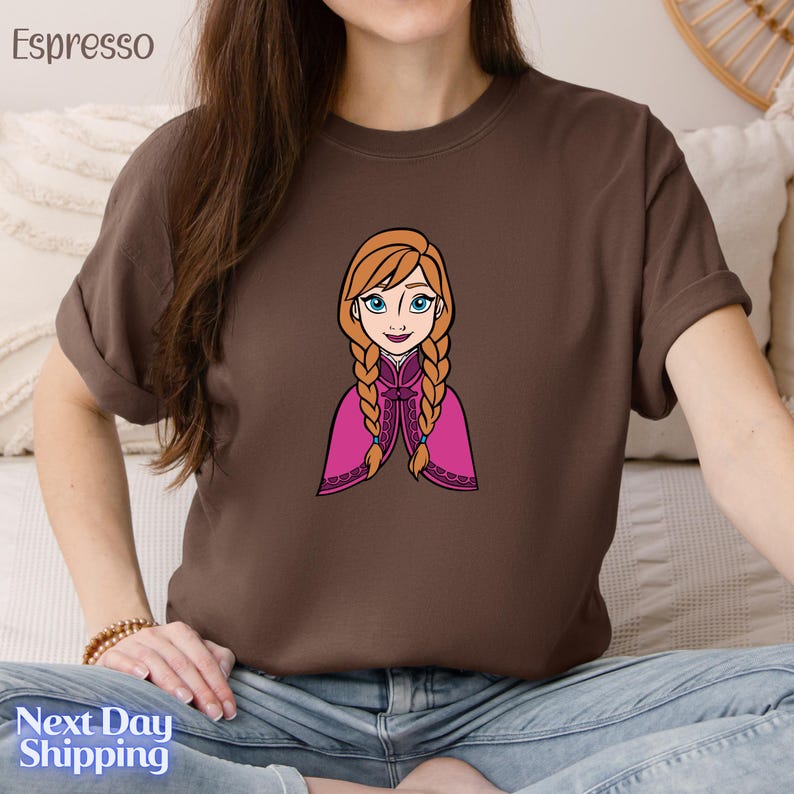 A woman wearing a brown t-shirt with a cartoon illustration of a young girl with braided hair and blue eyes.
