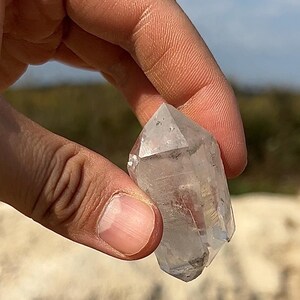 May include: A clear quartz crystal point held between fingers. The crystal is transparent with visible inclusions and facets. The background is blurred, suggesting an outdoor setting with natural light.