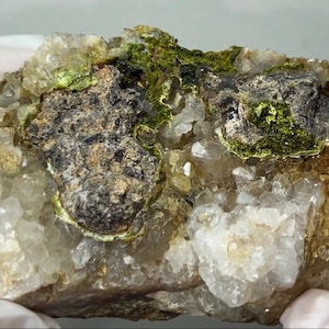 May include: A close-up of a mineral specimen featuring a mix of colors and textures. The rock has a base of clear quartz crystals, with patches of dark gray and green mineral deposits. The overall appearance is rough and natural.