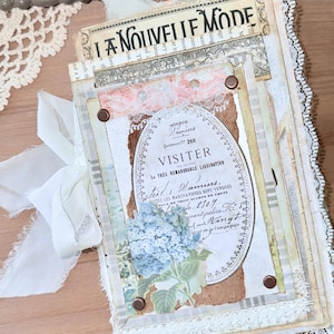 May include: A handmade journal with a vintage aesthetic. The cover features layered paper with lace, ribbon, and a label that reads "VISITER". A blue hydrangea illustration is visible. The journal is tied with a white ribbon.